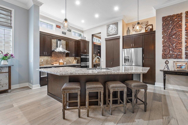 kitchen with dark brown cabinetry, wall chimney range hood, an island with sink, pendant lighting, and appliances with stainless steel finishes