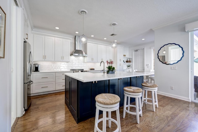 kitchen featuring hanging light fixtures, an island with sink, wall chimney exhaust hood, sink, and white cabinetry