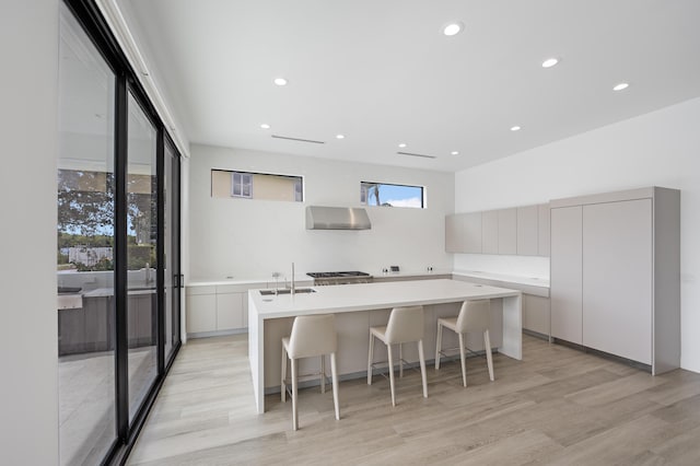 kitchen with a kitchen breakfast bar, wall chimney range hood, light wood-type flooring, an island with sink, and white cabinetry