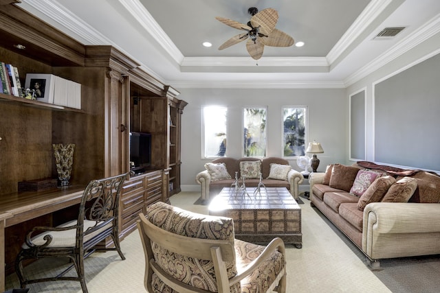 carpeted living room with a tray ceiling, ceiling fan, built in desk, and ornamental molding