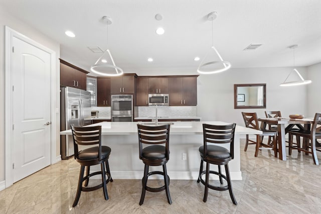 kitchen with decorative backsplash, appliances with stainless steel finishes, dark brown cabinetry, a kitchen island with sink, and decorative light fixtures