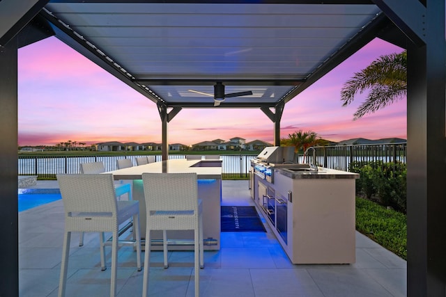 patio terrace at dusk featuring a water view, an outdoor kitchen, a grill, and ceiling fan