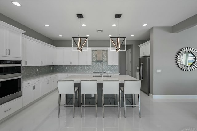 kitchen featuring appliances with stainless steel finishes, decorative light fixtures, white cabinets, wall chimney range hood, and a center island with sink