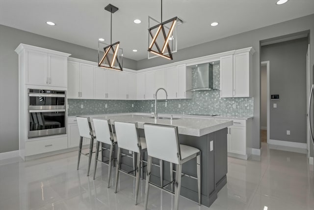 kitchen featuring white cabinetry, stainless steel double oven, a kitchen island with sink, and wall chimney range hood