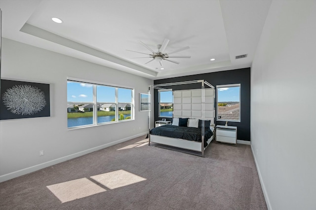 carpeted bedroom with multiple windows, a tray ceiling, ceiling fan, and a water view