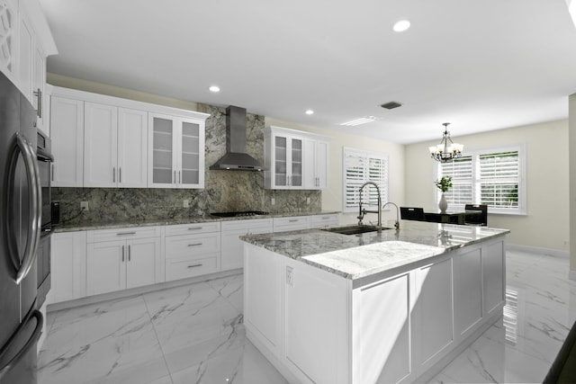 kitchen featuring light stone counters, wall chimney exhaust hood, sink, a center island with sink, and white cabinetry