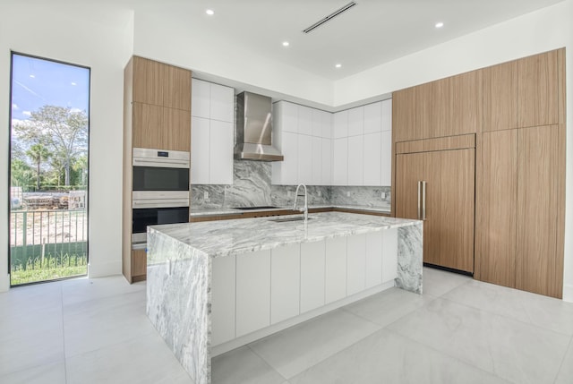 18416 Symphony Ct, Jupiter, FL 33458 - kitchen featuring wall chimney range hood, modern cabinets, double oven, a sink, and light stone countertops