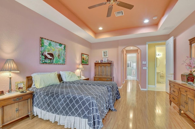 bedroom with connected bathroom, a tray ceiling, light hardwood / wood-style flooring, and ceiling fan