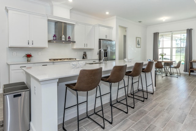 kitchen with sink, wall chimney exhaust hood, a kitchen island with sink, white cabinets, and appliances with stainless steel finishes