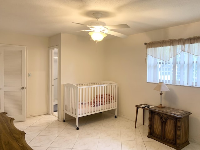 bedroom featuring ceiling fan, a textured ceiling, a closet, light tile patterned flooring, and a crib