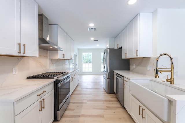 kitchen featuring white cabinets, wall chimney range hood, appliances with stainless steel finishes, and light hardwood / wood-style flooring