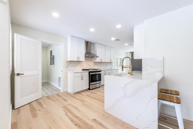 kitchen with wall chimney range hood, light stone countertops, stainless steel range oven, white cabinetry, and a breakfast bar area