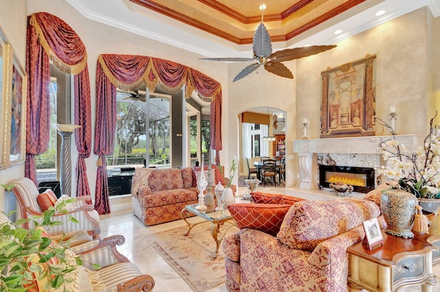 living room with a high end fireplace, a high ceiling, a raised ceiling, crown molding, and ceiling fan