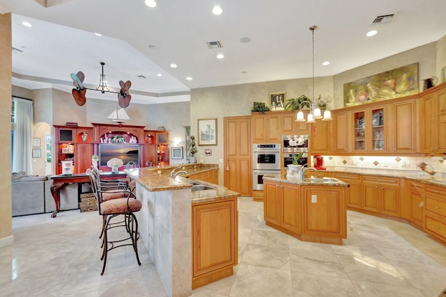 kitchen featuring stainless steel double oven, a kitchen island with sink, sink, pendant lighting, and a chandelier