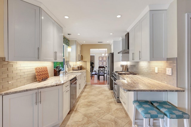 kitchen with backsplash, wall chimney range hood, light stone counters, stainless steel appliances, and a sink