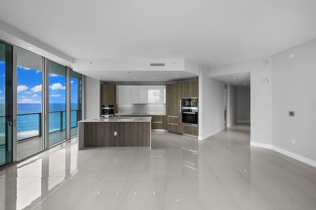 kitchen with white cabinetry, sink, a wall of windows, a center island with sink, and a water view