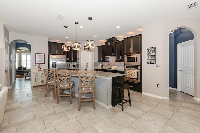 kitchen with dark brown cabinetry, a kitchen island with sink, decorative backsplash, appliances with stainless steel finishes, and custom exhaust hood