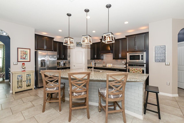 kitchen featuring appliances with stainless steel finishes, light stone counters, dark brown cabinetry, pendant lighting, and a center island with sink