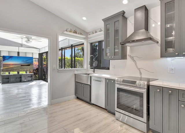 kitchen featuring gray cabinetry, stainless steel appliances, vaulted ceiling, ceiling fan, and wall chimney range hood