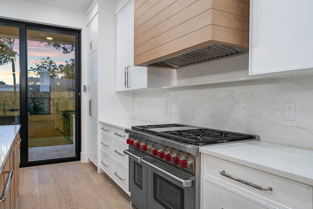 kitchen featuring light stone countertops, light wood-type flooring, premium range hood, range with two ovens, and white cabinetry