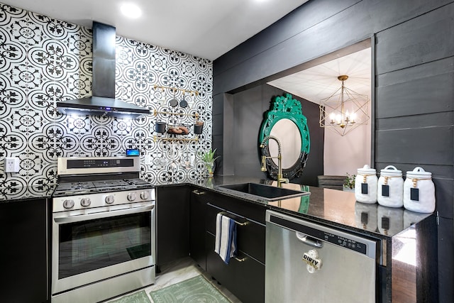 kitchen featuring wall chimney range hood, sink, appliances with stainless steel finishes, dark stone countertops, and a notable chandelier