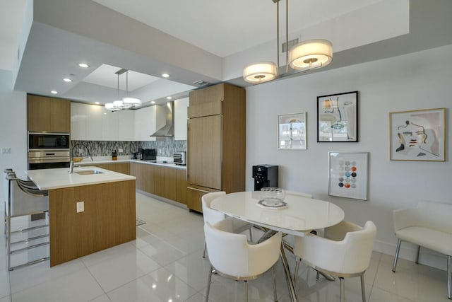 kitchen with backsplash, wall chimney exhaust hood, a kitchen island with sink, pendant lighting, and white cabinets