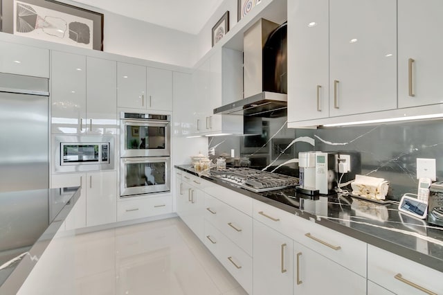 kitchen with wall chimney range hood, tasteful backsplash, white cabinets, built in appliances, and light tile patterned floors