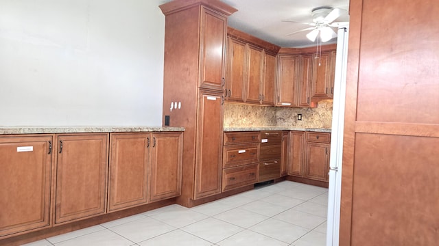 kitchen featuring light stone counters, light tile patterned floors, brown cabinetry, ceiling fan, and decorative backsplash