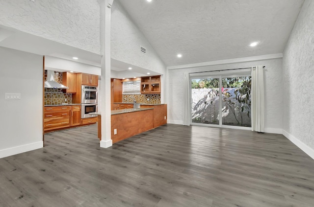 kitchen featuring dark wood-type flooring, wall chimney range hood, high vaulted ceiling, backsplash, and stainless steel double oven