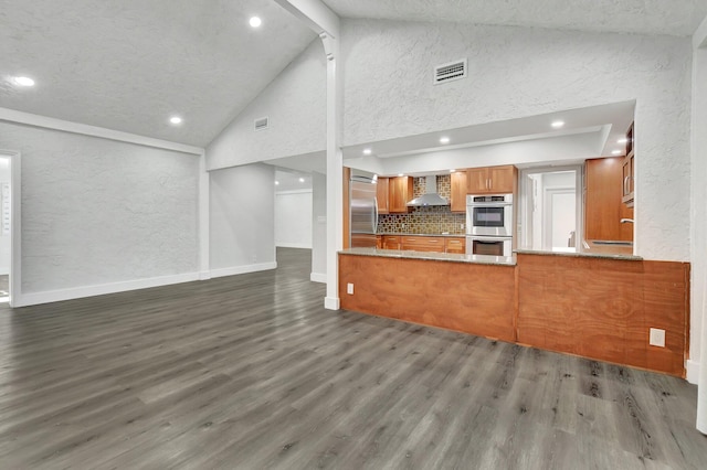 kitchen with stainless steel appliances, kitchen peninsula, dark hardwood / wood-style flooring, high vaulted ceiling, and wall chimney exhaust hood