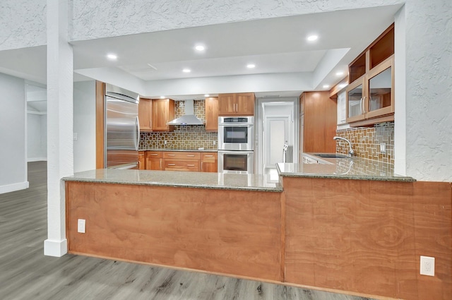 kitchen with kitchen peninsula, light stone countertops, wall chimney exhaust hood, and stainless steel appliances