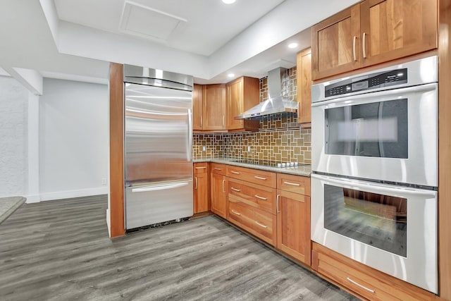 kitchen featuring light wood-style floors, appliances with stainless steel finishes, wall chimney range hood, decorative backsplash, and brown cabinets