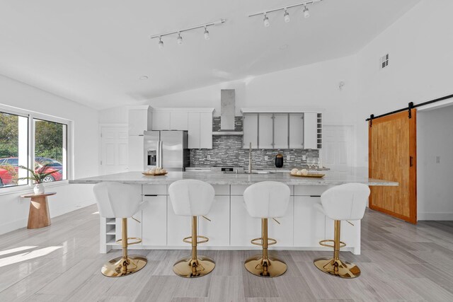 kitchen with decorative backsplash, stainless steel fridge, wall chimney exhaust hood, a barn door, and white cabinets