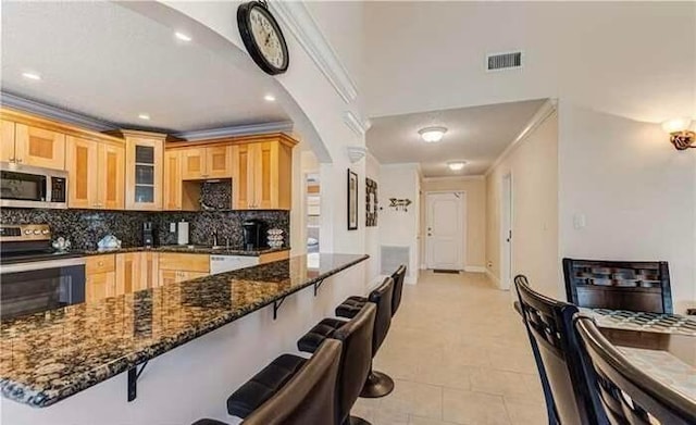 kitchen featuring decorative backsplash, dark stone countertops, appliances with stainless steel finishes, light tile patterned flooring, and a breakfast bar area