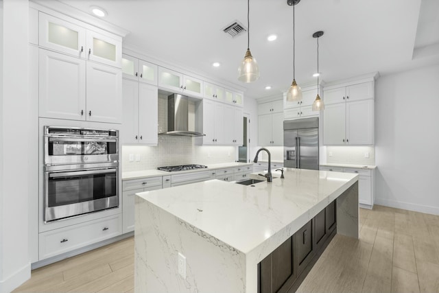 kitchen with white cabinets, an island with sink, stainless steel appliances, and wall chimney range hood