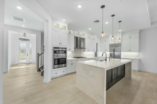 kitchen featuring stainless steel appliances, sink, wall chimney range hood, white cabinets, and an island with sink