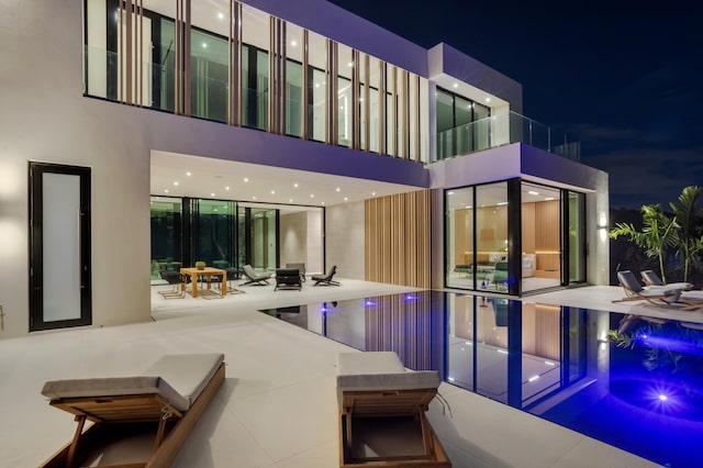 back of house at twilight featuring an infinity pool, a patio area, a balcony, and stucco siding