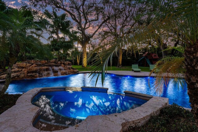 pool at dusk with pool water feature, a playground, and an in ground hot tub