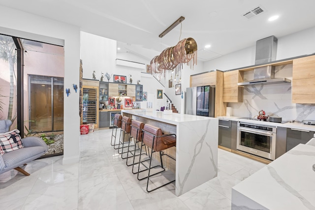 kitchen featuring a kitchen breakfast bar, hanging light fixtures, wall chimney exhaust hood, light stone countertops, and stainless steel appliances