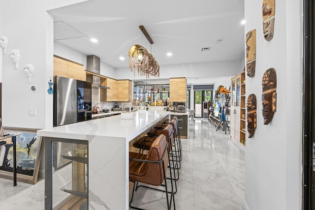 kitchen with light stone countertops, wall chimney range hood, pendant lighting, light brown cabinets, and a kitchen island