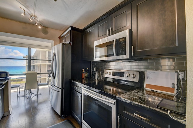 kitchen featuring appliances with stainless steel finishes, backsplash, dark brown cabinets, a water view, and dark stone countertops