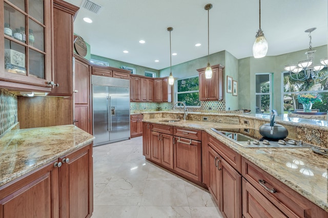 kitchen with backsplash, sink, hanging light fixtures, light stone countertops, and stainless steel built in refrigerator