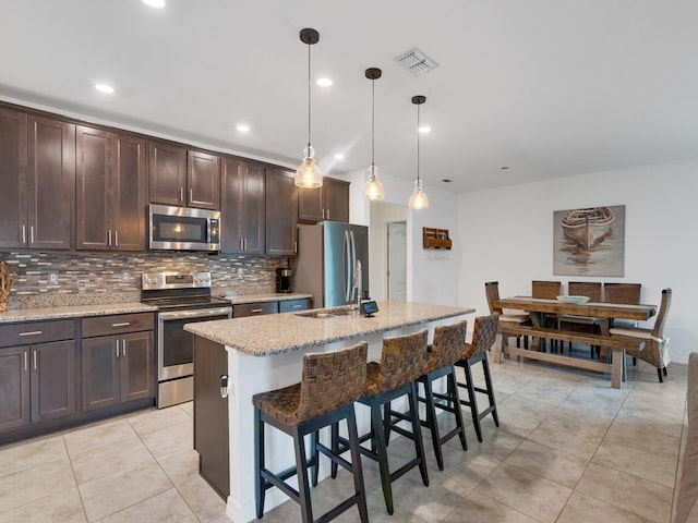 kitchen with light stone countertops, appliances with stainless steel finishes, a kitchen breakfast bar, a center island with sink, and hanging light fixtures