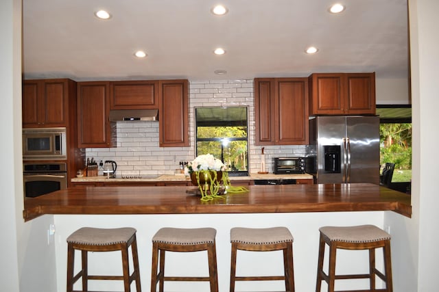 kitchen featuring exhaust hood, wooden counters, decorative backsplash, a kitchen bar, and stainless steel appliances