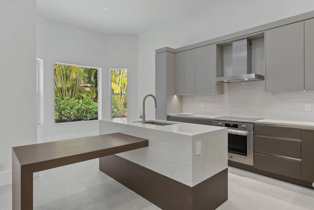 kitchen featuring gray cabinetry, sink, wall chimney range hood, oven, and an island with sink