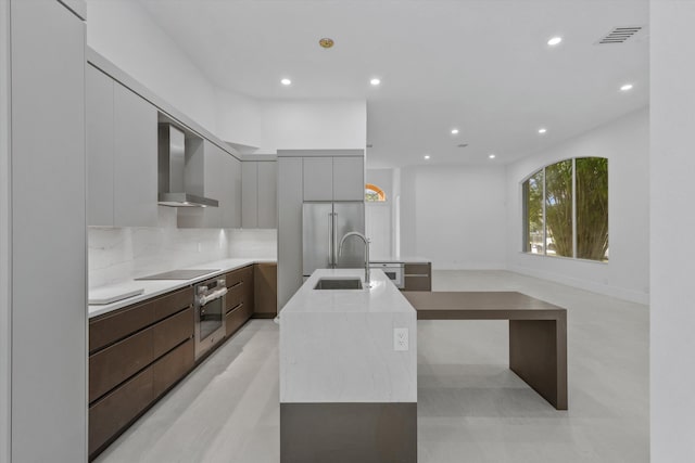 kitchen with appliances with stainless steel finishes, tasteful backsplash, wall chimney exhaust hood, sink, and a center island with sink