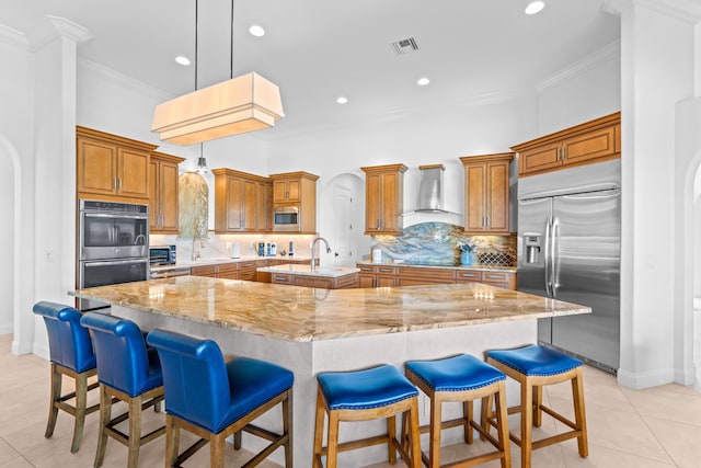 kitchen featuring a large island with sink, built in appliances, wall chimney exhaust hood, light stone countertops, and decorative light fixtures