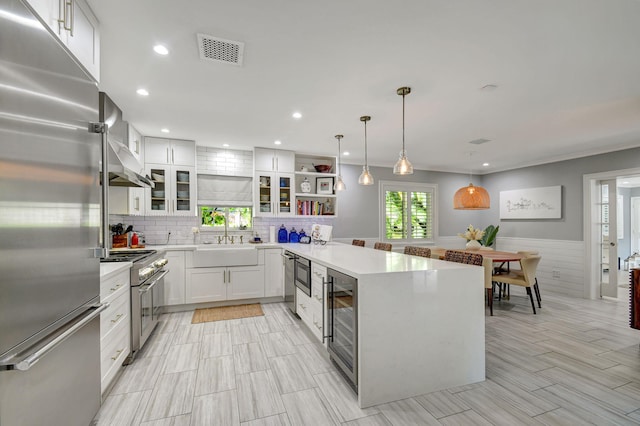 kitchen featuring wall chimney range hood, sink, built in appliances, decorative light fixtures, and white cabinetry