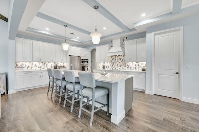 kitchen featuring arched walkways, a tray ceiling, stainless steel appliances, light wood-style floors, and premium range hood