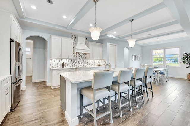 kitchen with pendant lighting, stainless steel refrigerator with ice dispenser, premium range hood, and a kitchen island with sink